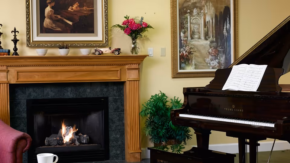 A cozy interior room featuring a lit fireplace with a wooden mantel decorated with small bowls, a toy car, and a vase of pink flowers. To the right, there is a grand piano with sheet music on the stand, and two framed paintings hang on the yellow walls. A green plant is placed between the fireplace and piano, and part of a red armchair with a white cup on the arm is visible on the left.