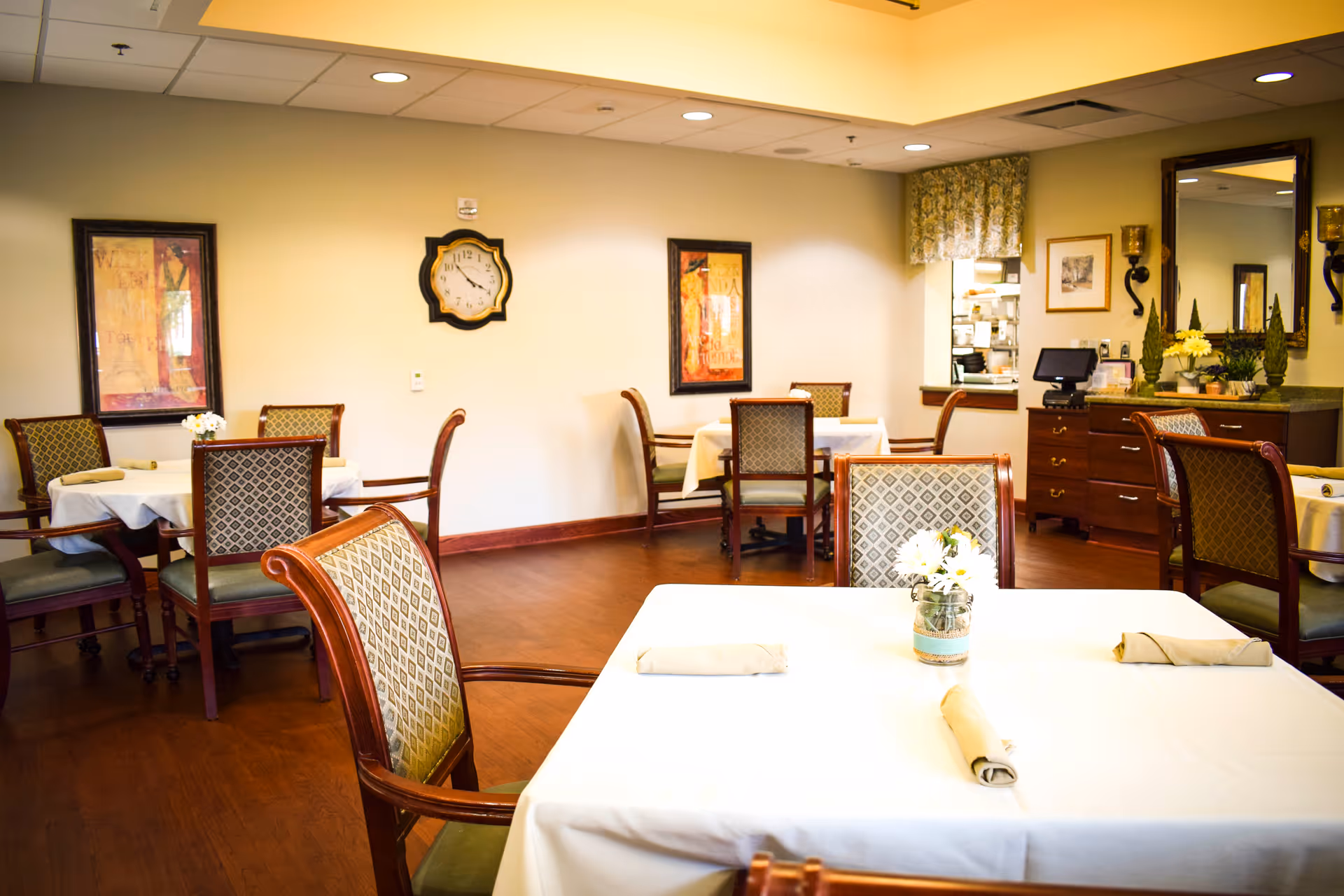 A dining room with several tables covered in white tablecloths, each set with rolled napkins and small floral centerpieces. The room has wooden chairs with patterned upholstery, framed artwork on the walls, a clock, and a sideboard with a mirror and decorative items.