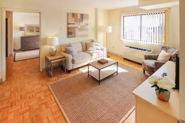 A bright and cozy living room with a beige sofa, patterned armchair, wooden coffee table on a large woven rug, side table with a lamp, and a large window with vertical blinds letting in natural light. A doorway leads to a bedroom with a dresser and mirror visible.