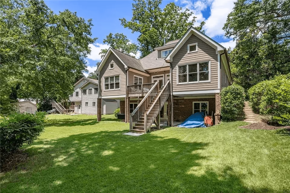Exterior view of a two-story house with beige siding and a brick foundation, featuring a wooden staircase leading to a small deck. The house is surrounded by a well-maintained green lawn, trees, and bushes under a blue sky with some clouds.