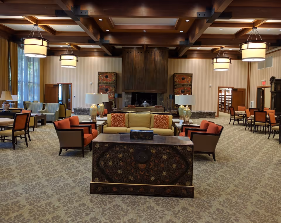 A spacious and elegant living room area with a high wooden beamed ceiling and large hanging light fixtures. The room features a central seating arrangement with a yellow sofa, red armchairs, and decorative pillows. There are side tables with lamps, patterned carpet, and large windows with curtains on the left side. In the background, there is a large wooden fireplace flanked by decorative cabinets. Additional seating and tables are arranged around the room.