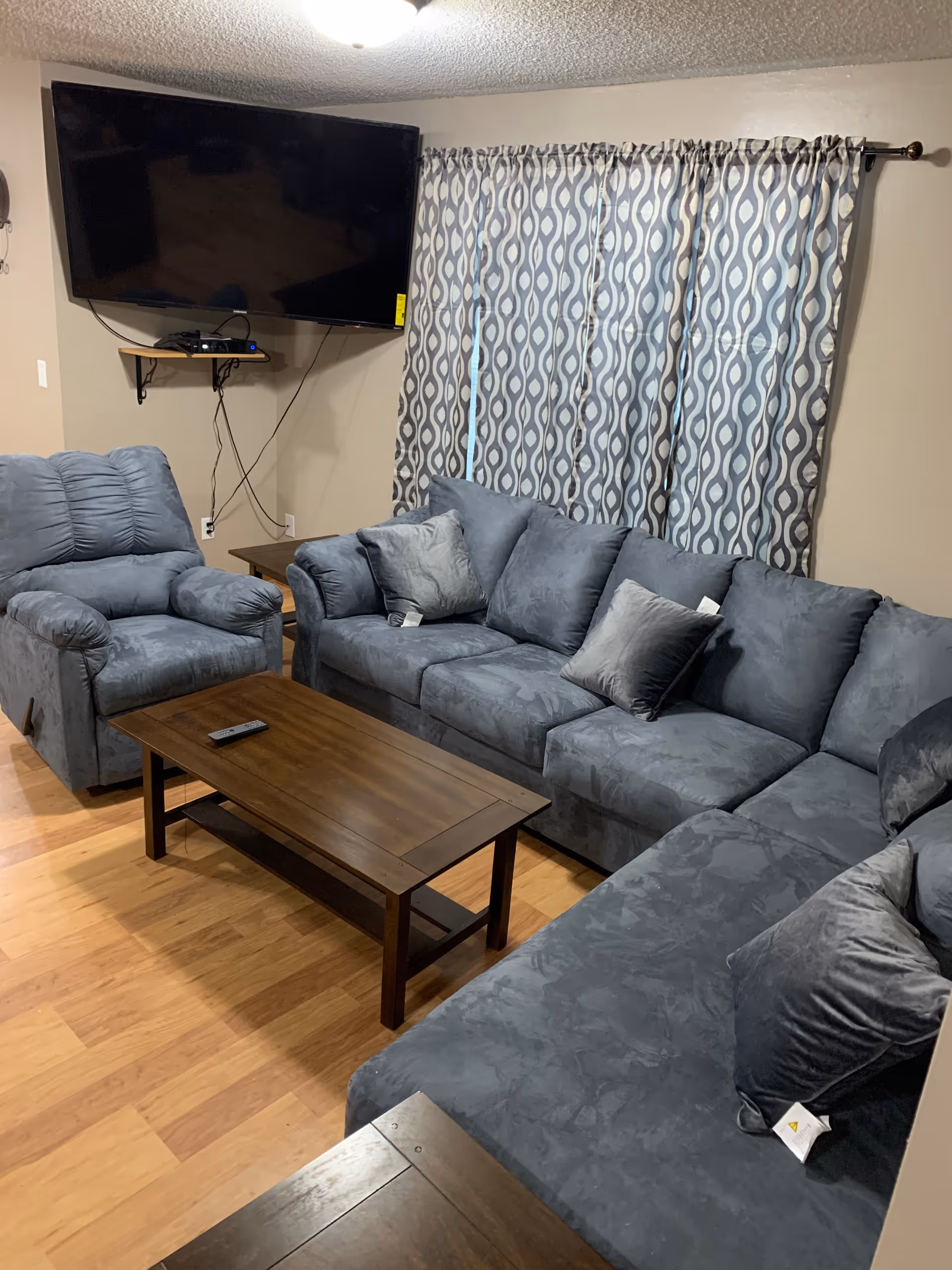 Living room with blue-gray sectional sofa and armchair, wooden coffee table, wall-mounted TV, and patterned curtains.