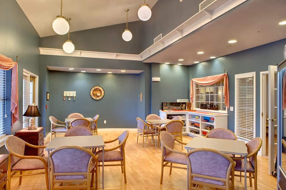 A well-lit common area with several tables and chairs arranged for group seating. The walls are painted blue with decorative items including a clock and a sign. There are windows with red and white checkered curtains, a lamp on a small table, and shelves with board games and other items. The ceiling has hanging globe lights and recessed lighting.