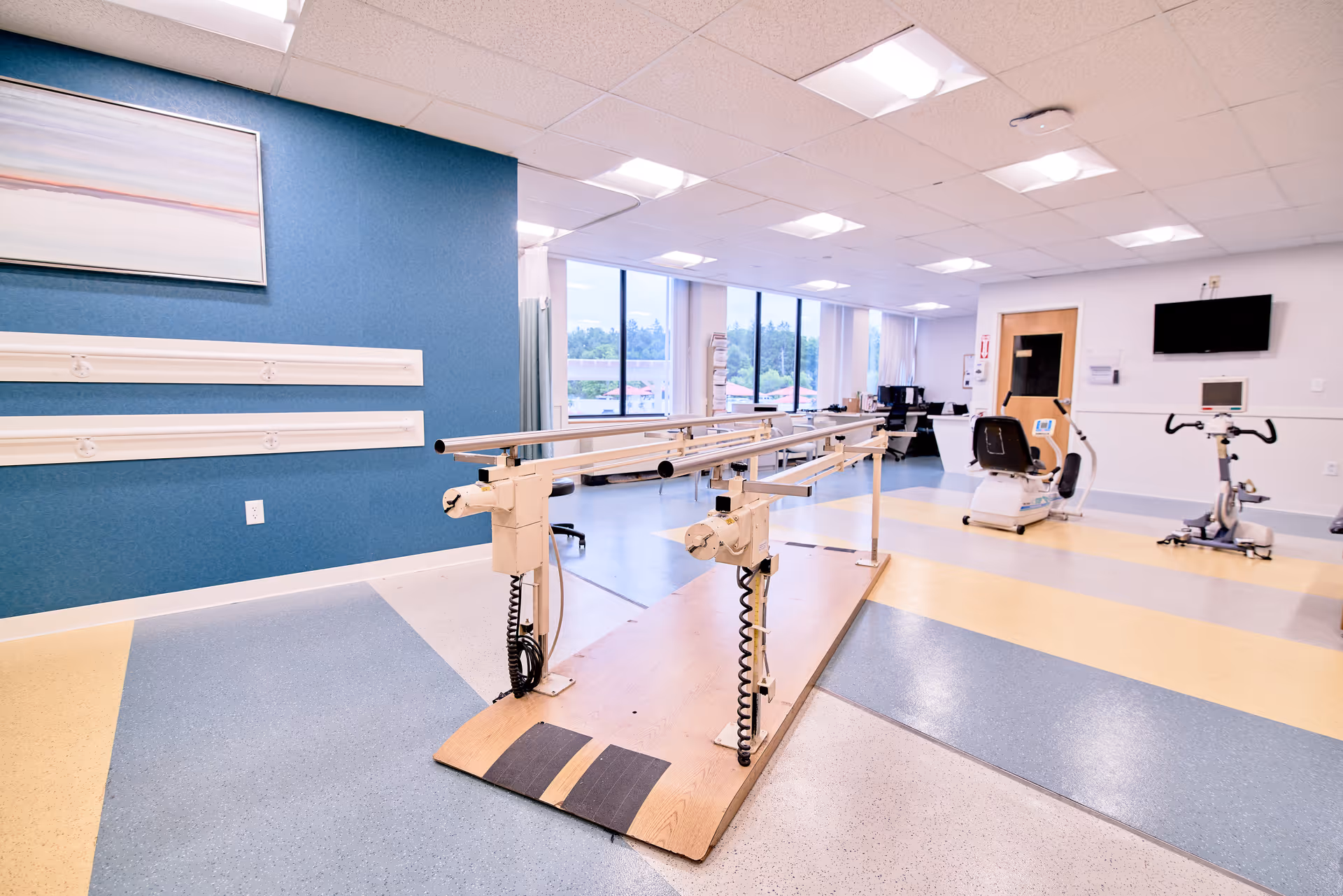 A bright physical therapy room with parallel bars for walking exercises in the center, exercise bikes on the right, large windows letting in natural light, and a blue accent wall with a framed abstract painting.