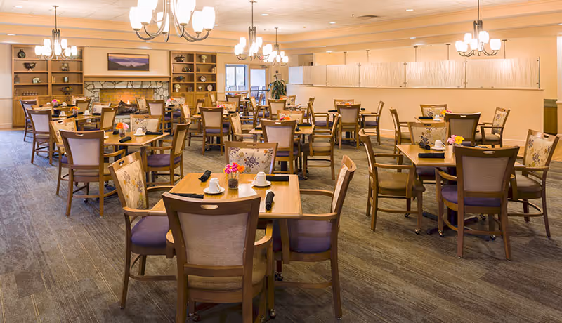 A spacious dining room with multiple wooden tables and chairs arranged neatly. Each table is set with white cups, napkins, and small flower vases. The room features warm lighting from chandeliers, a carpeted floor, and a stone fireplace with a painting above it. Large windows allow natural light to enter the space.