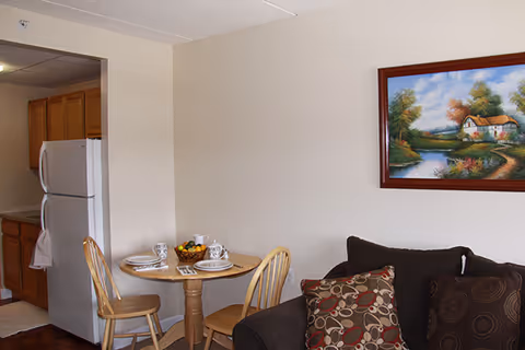 A small dining area with a round wooden table set for two, featuring plates, cups, and a bowl of fruit. Two wooden chairs are placed around the table. To the left, a kitchen area with wooden cabinets and a white refrigerator is visible. On the right, part of a dark brown couch with patterned cushions is seen. A framed painting of a countryside scene with a house, trees, and a river hangs on the wall above the couch.
