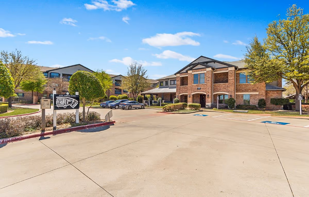 Exterior view of The Cesera 55+ Apartments showing a large paved parking area with several parked cars, a site directory sign, and two-story brick buildings surrounded by trees and landscaping under a blue sky with scattered clouds.