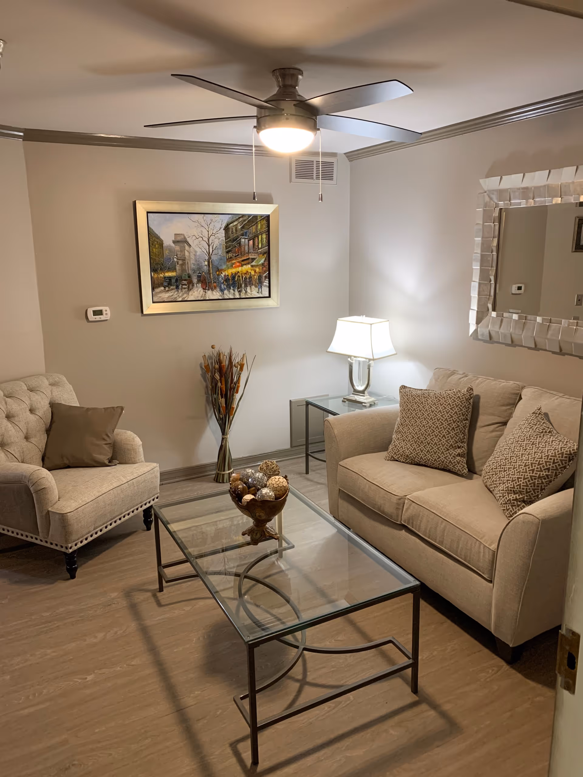 A cozy living room with a beige loveseat and armchair, both with patterned cushions. A glass coffee table with decorative items sits in the center on a wooden floor. A table lamp on a glass side table illuminates the room. A large mirror with a textured frame hangs on the wall above the loveseat, and a framed painting of a street scene is on the adjacent wall. A ceiling fan with a light is mounted on the ceiling.