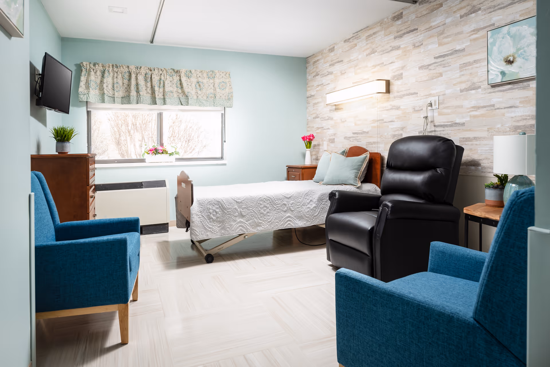 A bright and clean senior living bedroom with a single bed covered in a white quilt, two blue armchairs, a black recliner chair, a wooden dresser with a small plant, a window with floral curtains, and a side table with a lamp and flowers. The walls are painted light blue and one wall has a textured stone-like design.