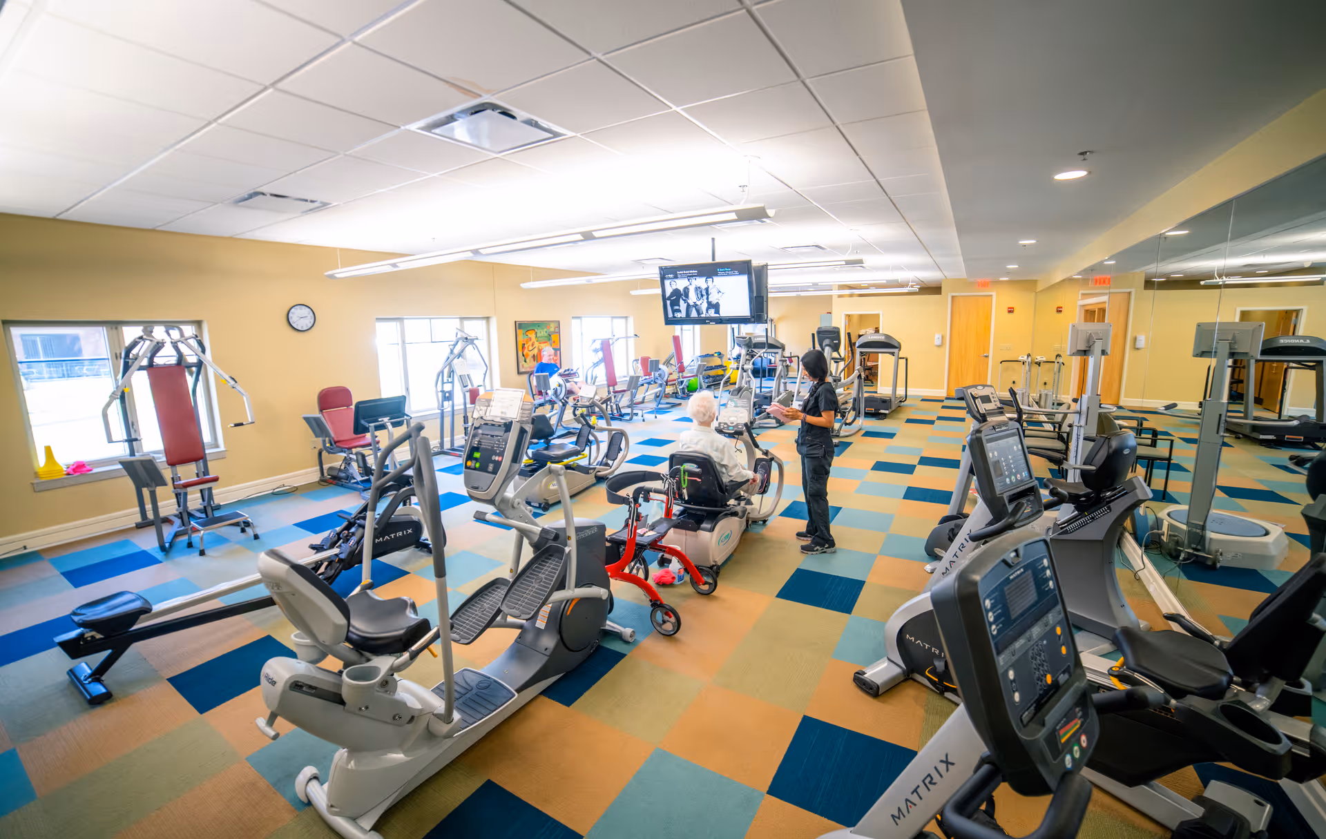 A brightly lit fitness room with various exercise equipment including stationary bikes, treadmills, and weight machines. An elderly person is seated on a recumbent bike while a staff member stands nearby. The room has large windows, a colorful checkered carpet, and a large mirror on one wall. A television is mounted from the ceiling in the center of the room.