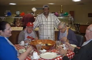 A group of elderly people sitting around a table with a red and white checkered tablecloth, enjoying a meal together. A man in a plaid shirt stands behind them smiling. The setting appears to be a communal dining area with kitchen shelves and utensils visible in the background.