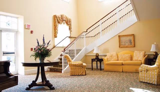 A spacious and well-lit living room area with a yellow sofa, two matching armchairs, a small side table with a lamp, a round wooden table with a flower arrangement, a staircase with white railings, and a piano near the entrance door.
