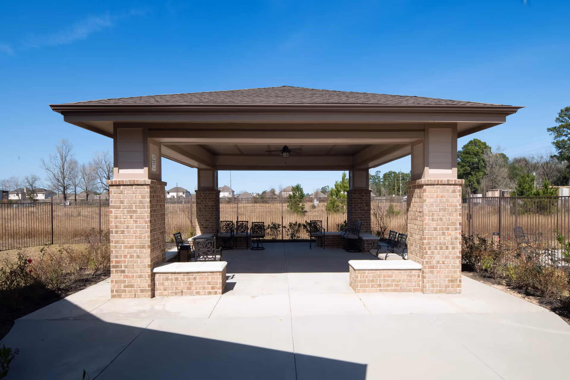 Covered brick pavilion with benches and chairs in an outdoor courtyard under a clear blue sky.