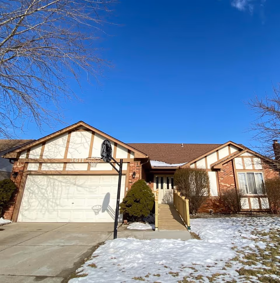 Front exterior view of a single-story brick house with a two-car garage, a basketball hoop in the driveway, a wooden ramp leading to the front door, and some snow on the ground and roof under a clear blue sky.