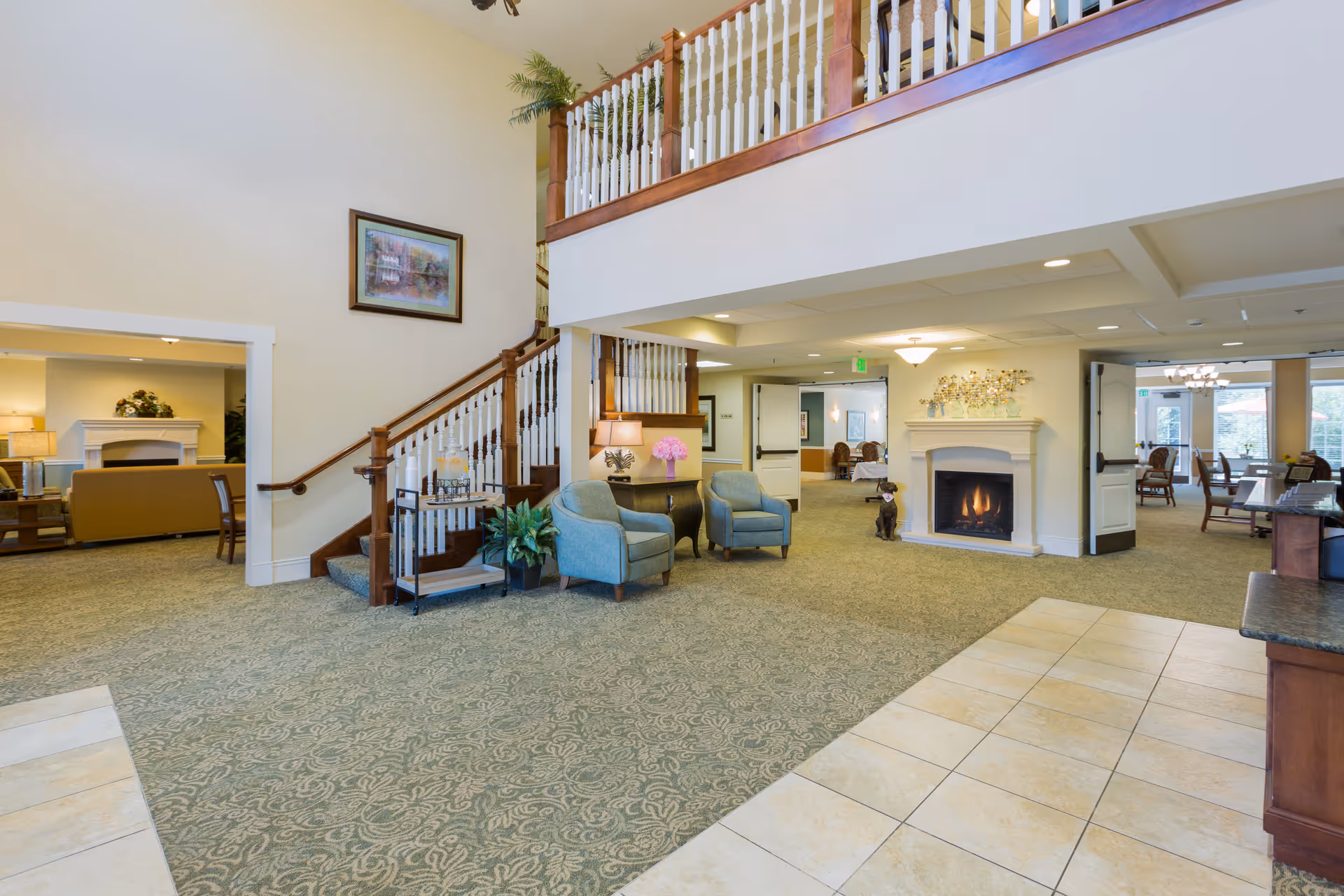 Spacious and well-lit common area in an assisted living facility featuring a staircase with wooden railings, two blue armchairs with a small table and lamp between them, a fireplace with a decorative piece above it, and a dining area visible in the background. The floor is carpeted with a patterned design, and the ceiling has recessed lighting.