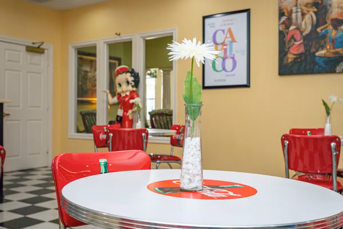 A retro-style dining area with a round white table featuring a Coca-Cola logo, surrounded by red chairs. A tall glass vase with a white flower and green leaves sits on the table. In the background, there is a life-sized Betty Boop figure dressed in red, a checkered black and white floor, and framed artwork on yellow walls.