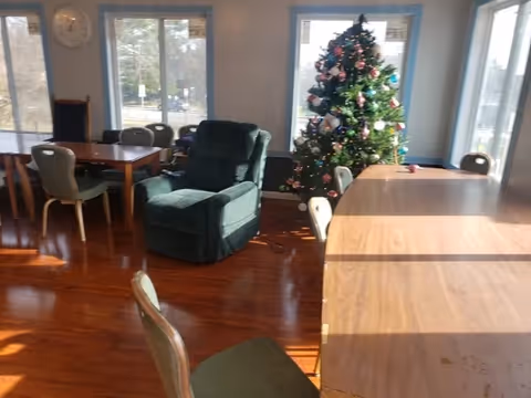 Sunlit common room with tables and chairs, a green recliner, and a decorated Christmas tree by the windows.