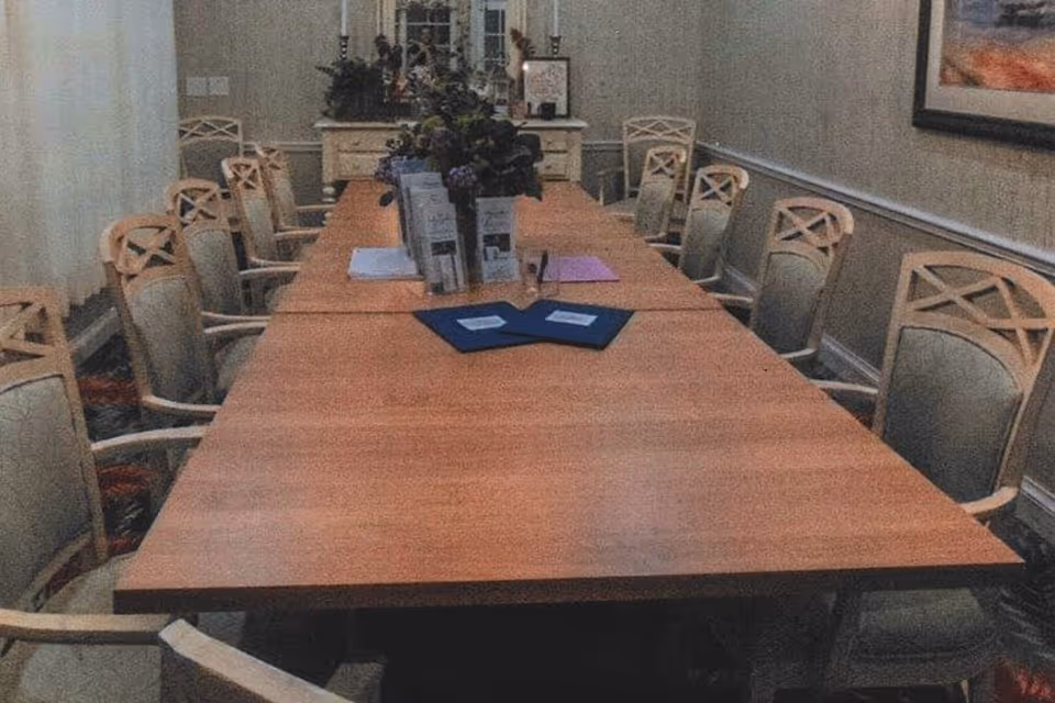 A long wooden conference or dining table surrounded by multiple beige cushioned chairs with armrests in a well-lit room. The table has some documents, folders, and a flower arrangement in the center. The room has light-colored walls with a framed picture hanging on one side and a cabinet with decorative items at the far end.