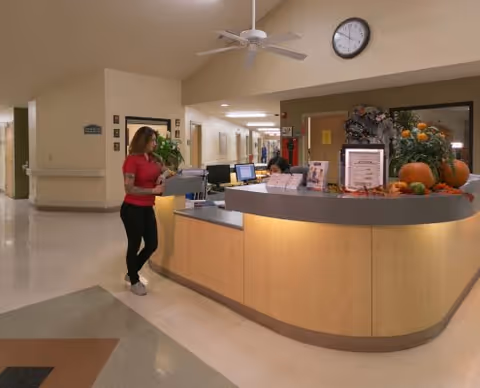 A woman in a red shirt standing at a curved reception desk in a healthcare facility. Behind the desk, a staff member is seated working on a computer. The desk is decorated with autumn-themed items including pumpkins and fall leaves. A clock is mounted on the wall above the desk, and a hallway extends into the background.