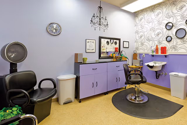 Interior of a hair salon area in a senior living facility with a black salon chair on a black mat, a purple cabinet with various items on top, a wall-mounted sink, and decorative wallpaper with circular mirrors on the wall.
