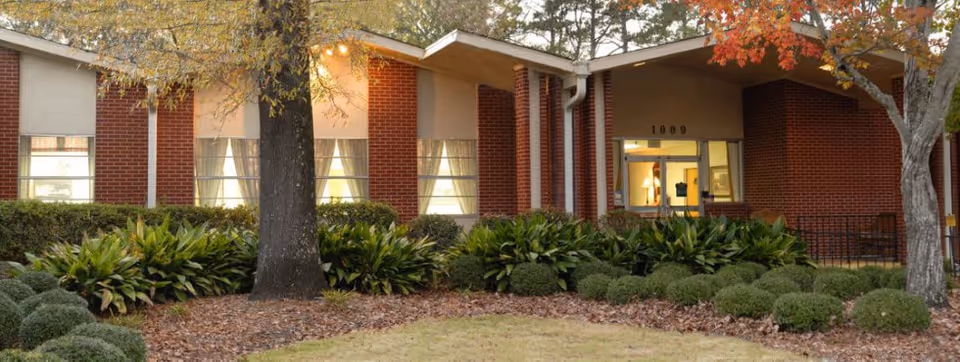 Exterior view of a single-story brick building with large windows and a glass entrance door. The building is surrounded by landscaped bushes, trees with autumn leaves, and a grassy area in front.