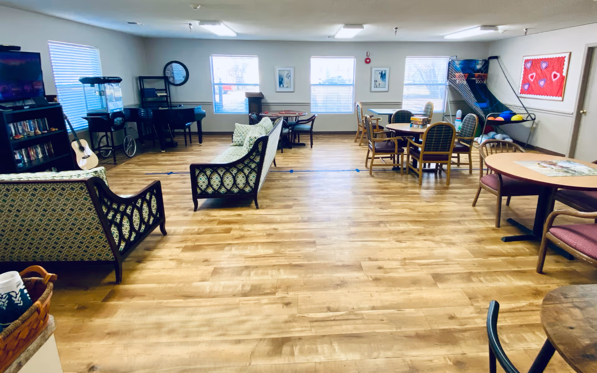 A spacious common area with wooden flooring featuring multiple seating arrangements including patterned sofas and round tables with chairs. The room has several windows letting in natural light, a television mounted on the wall, a popcorn machine, a guitar, a piano, and a basketball arcade game. The walls are decorated with framed pictures and a bulletin board with heart decorations.
