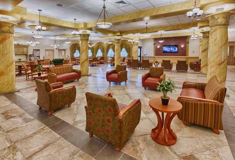 Spacious senior living facility lobby lounge with marble floors, yellow columns, and red upholstered chairs.