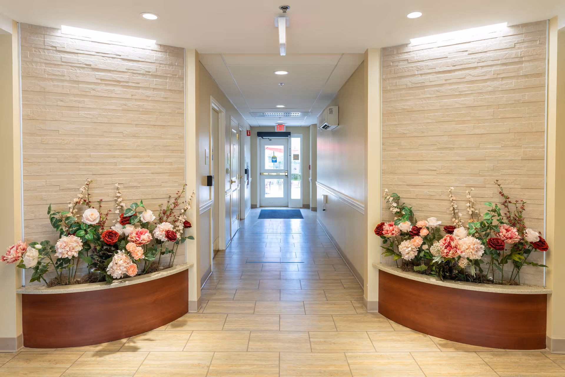 A well-lit hallway with beige tiled flooring and cream-colored walls. On either side of the hallway entrance, there are two curved wooden planters filled with artificial flowers in shades of pink, red, and white. The hallway leads to a glass door with an exit sign above it, and there are several doors along the left side of the hallway.