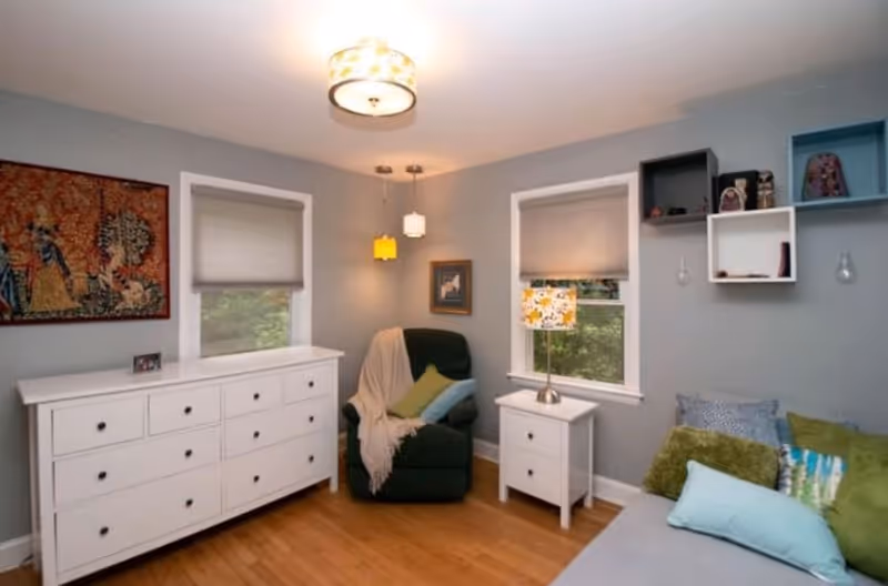 A cozy living room with light gray walls and wooden flooring. The room features a dark green armchair with a beige throw and colorful pillows, a white dresser with multiple drawers, a white side table with a floral lamp, two windows with gray blinds, and wall-mounted shelves with decorative items. A framed artwork hangs on the wall above the armchair.