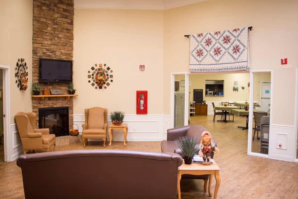 A cozy living room area in a senior living facility with a stone fireplace, a wall-mounted TV, two beige armchairs, a brown leather sofa, and a wooden side table with decorative plants and a small scarecrow figure. The room has light-colored walls and wood flooring, with an open doorway leading to a dining area with tables and chairs.