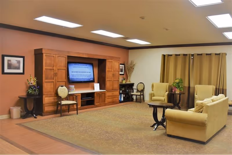 A spacious living room area with beige armchairs and a sofa arranged around small dark wood tables. A large wooden entertainment center with a flat-screen TV is mounted on a warm-toned wall. The room has a large carpet, framed pictures on the walls, and curtains covering the windows.