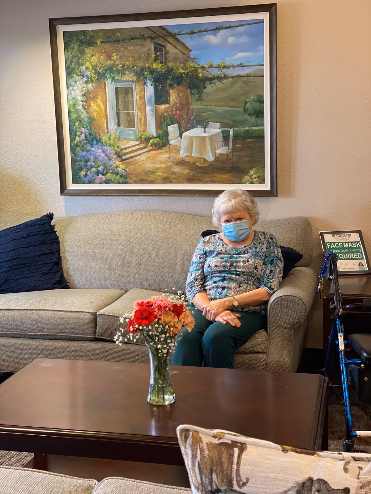 An older woman wearing a face mask sits on a couch in a lounge area with a coffee table holding a vase of flowers and a large framed painting on the wall.