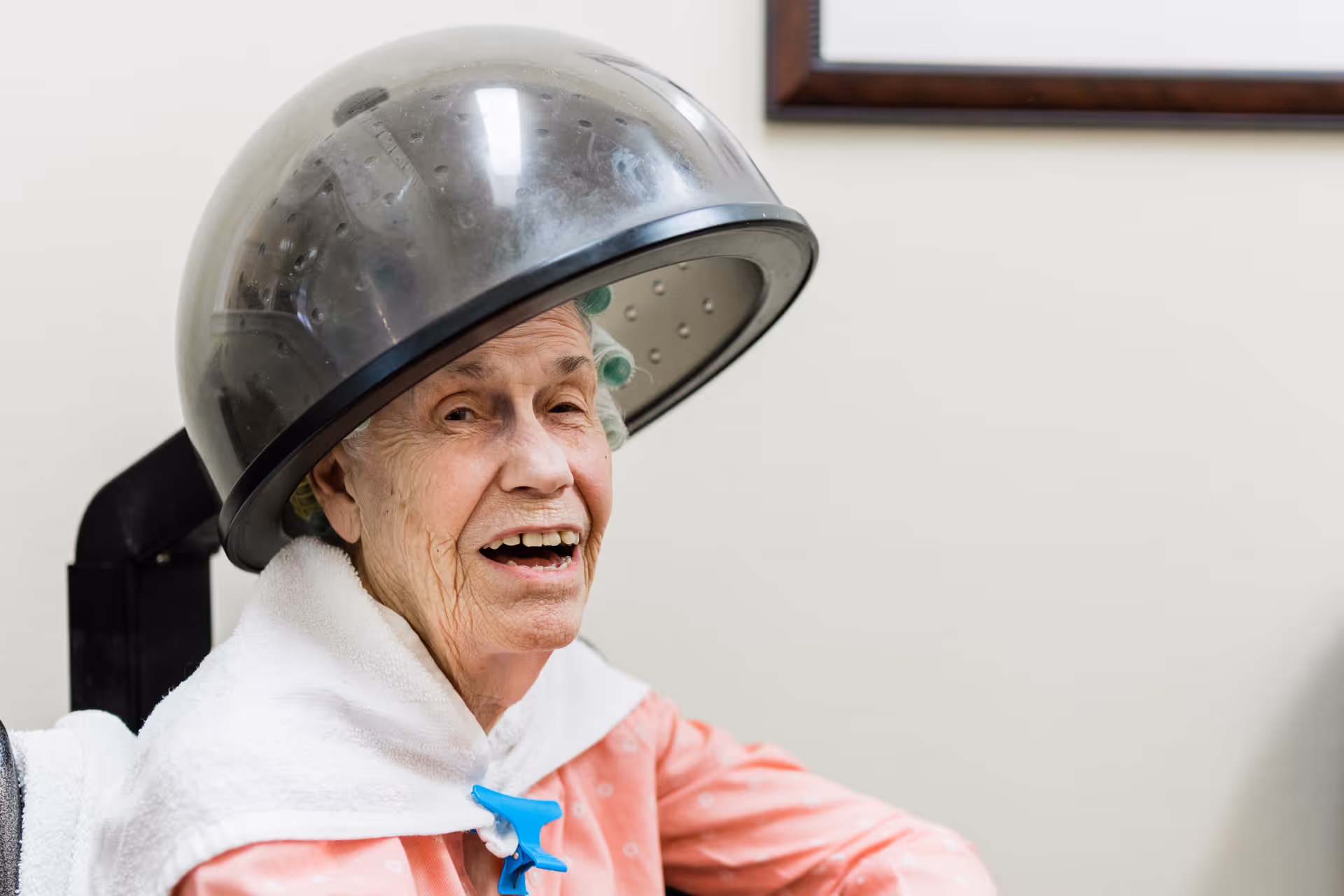 An elderly woman sitting under a hair dryer in a salon setting, wearing a pink robe and a white towel around her shoulders, smiling with her mouth slightly open.