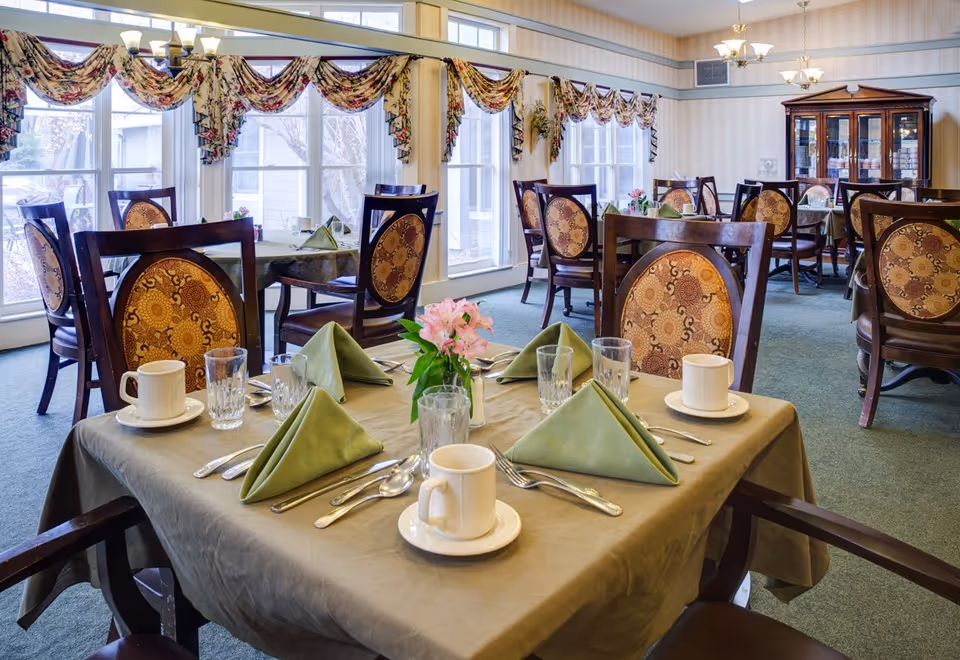 Dining room with multiple set tables featuring green napkins, cups, glassware and a floral centerpiece beneath large windows.
