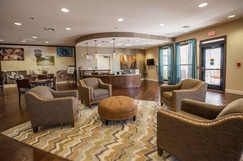 A spacious senior living facility common area with four cushioned armchairs arranged around a round leather ottoman on a patterned rug. The room features wood flooring, a reception desk in the background, wall art, and large windows with teal curtains allowing natural light. There is a door leading outside on the right side.
