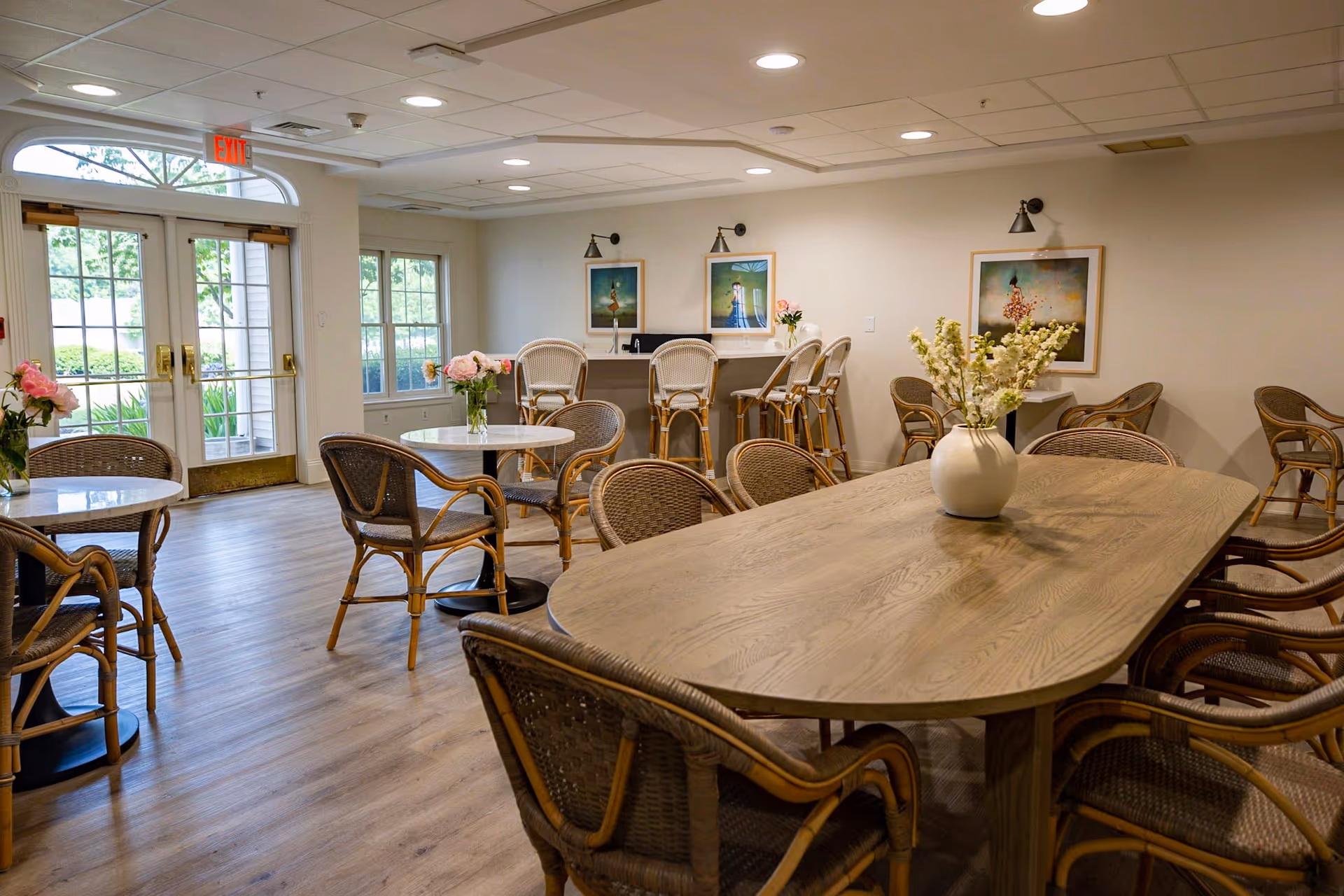 A bright and inviting dining area with multiple tables and wicker chairs. There is a large wooden table with a white vase of flowers in the foreground, smaller round tables with flower vases, and a counter with high chairs against the wall. The room has large windows and glass doors letting in natural light, light-colored walls, and framed artwork on the walls.