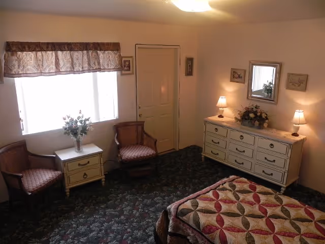 A bedroom with a bed covered in a patterned quilt, two wooden chairs with cushions, a small white nightstand with a vase of flowers, a white dresser with two lamps and a floral arrangement, a mirror and framed pictures on the walls, and a window with a valance letting in natural light.