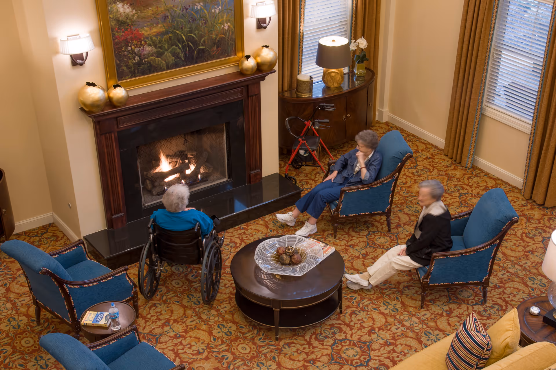 Three elderly women sitting and relaxing in a cozy living room with a lit fireplace. One woman is in a wheelchair facing the fireplace, while the other two are seated in blue armchairs. The room has a patterned carpet, a round coffee table with decorative items, and a side table with a water bottle and book. There are windows with curtains and a lamp on a wooden cabinet in the background.