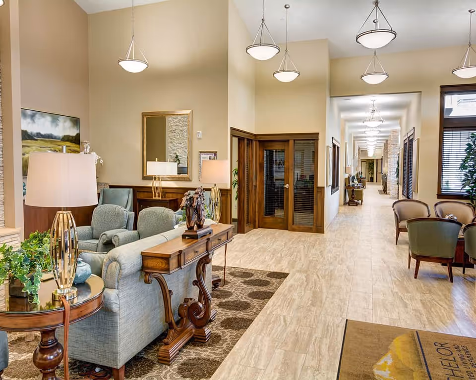 A bright assisted-living lobby with seating, decorative tables, and a long hallway lined with pendant lights.