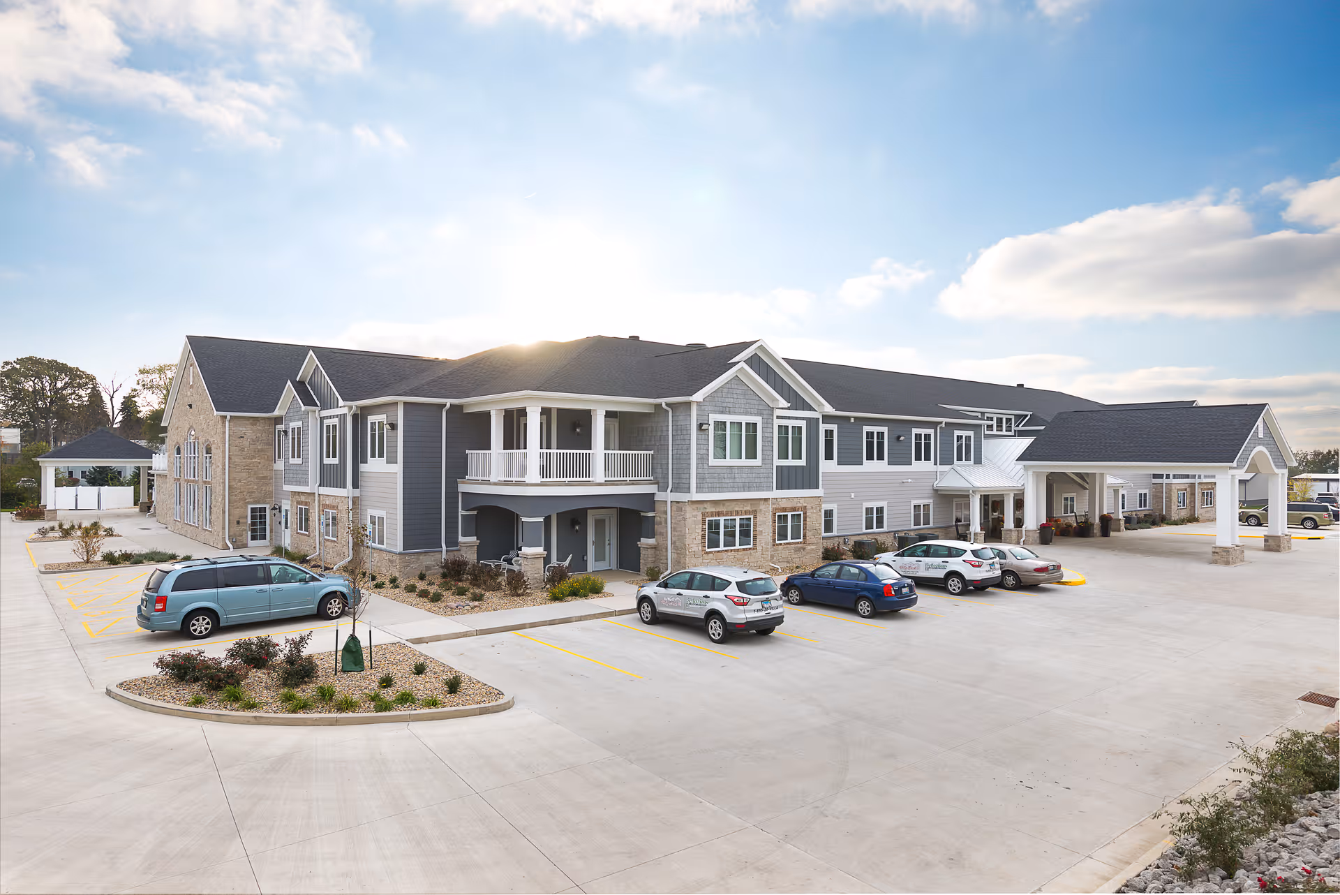 Exterior view of a modern assisted living and memory care facility building with a large parking lot in front. The building has a combination of stone and siding exterior with multiple windows and a covered entrance. Several cars are parked in the lot under a partly cloudy sky.
