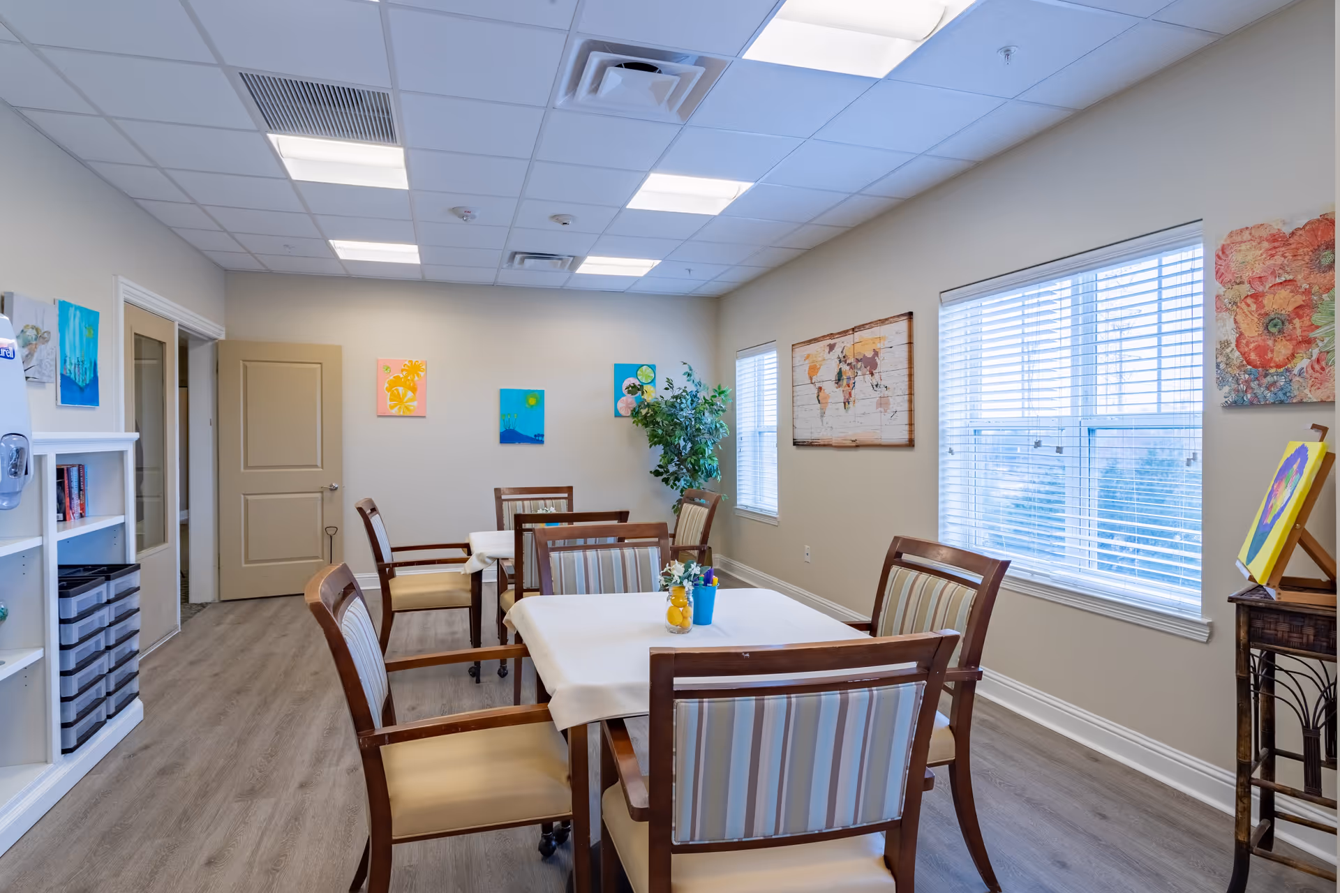 A bright and clean dining room with two tables covered with white tablecloths, surrounded by wooden chairs with striped cushions. The room has light-colored walls adorned with colorful paintings and a world map. Large windows with white blinds let in natural light, and a potted plant is placed in the corner. The floor is wood laminate, and the ceiling has recessed lighting panels.