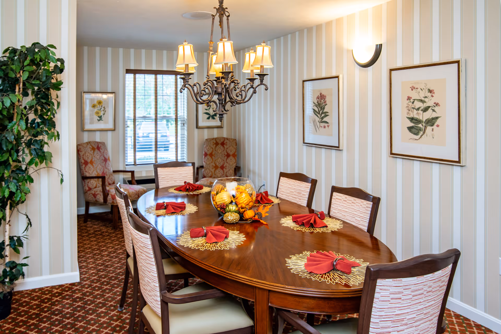 A dining room with a large oval wooden table set with six chairs. The table is decorated with red napkins on gold placemats and a centerpiece featuring autumn-themed decorations. The room has striped wallpaper, framed botanical prints on the walls, a chandelier overhead, and two upholstered armchairs near a window with blinds. A green potted plant is visible on the left side.