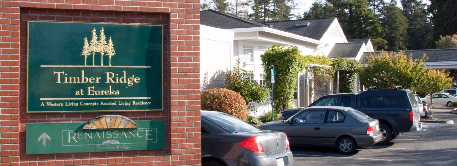 Exterior view of Timber Ridge at Eureka assisted living residence showing a green sign on a brick wall with the facility name and a parking lot with several parked cars and greenery in the background.