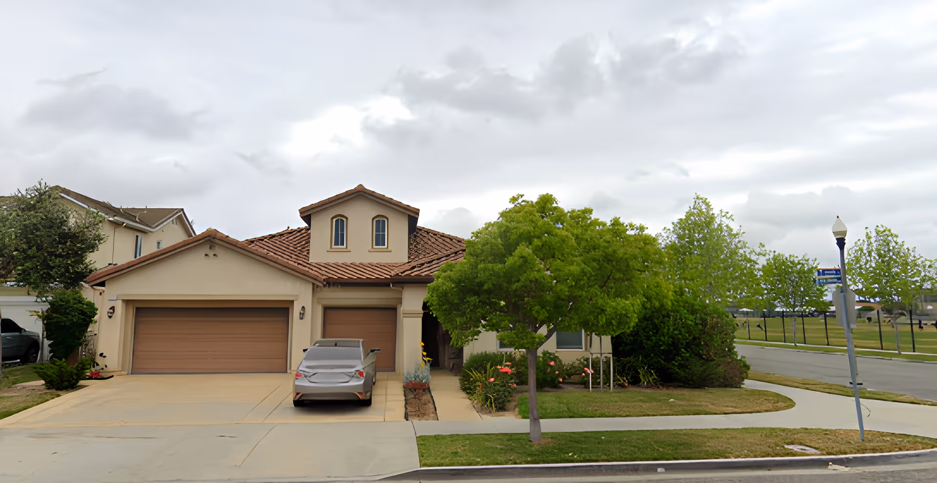 Front exterior view of a beige two-story house with a brown tiled roof, a three-car garage with closed doors, a parked car in the driveway, a small tree, and a sidewalk in front. The sky is cloudy and there is a street lamp and some greenery on the right side.