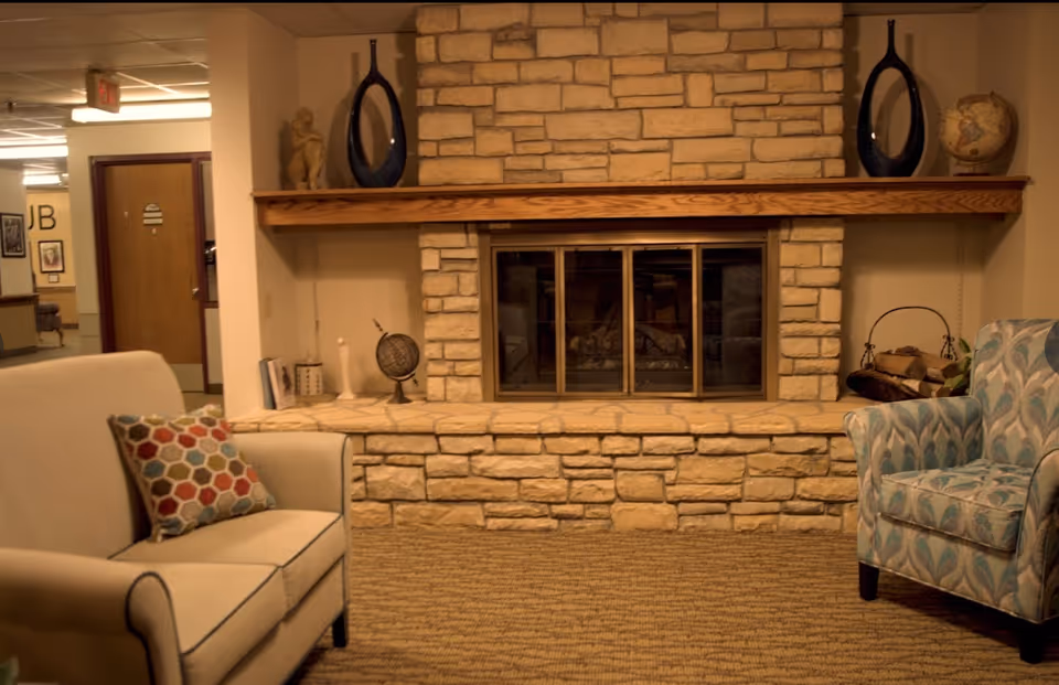 Cozy seating area with upholstered chairs and a stone fireplace hearth in a senior living common room.