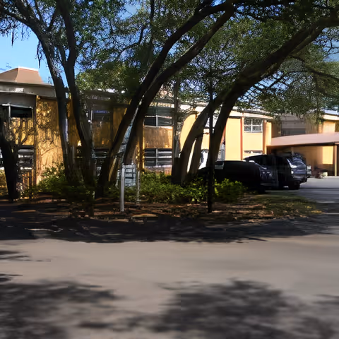 Exterior view of a senior living facility building partially shaded by large trees with several parked cars near the entrance under a covered area.