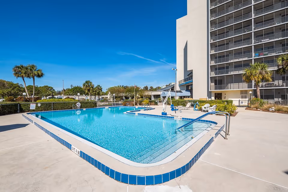 Outdoor swimming pool with clear blue water surrounded by a concrete deck, palm trees, and a multi-story building in the background under a clear blue sky.