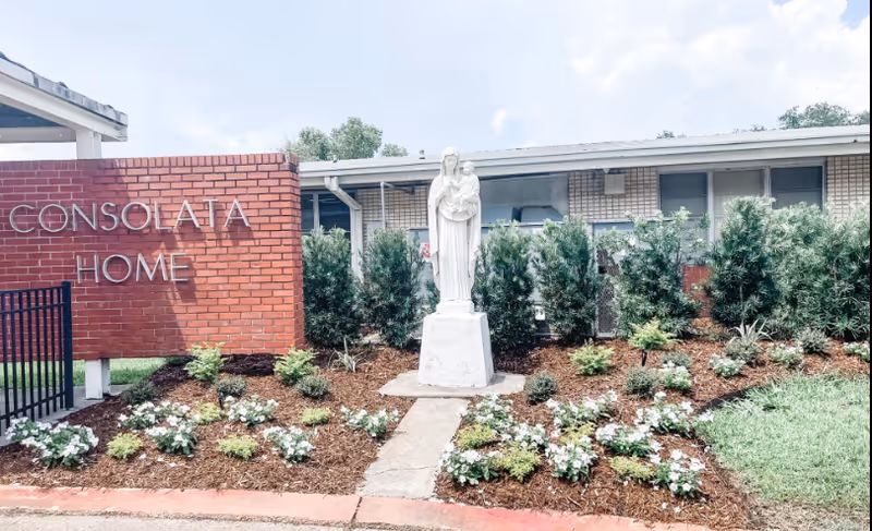 Outdoor garden area in front of a building with a red brick sign that reads 'Consolata Home'. In the center of the garden is a white statue of a person holding a child, surrounded by small plants and flowers. The building behind has windows and bushes along the wall.