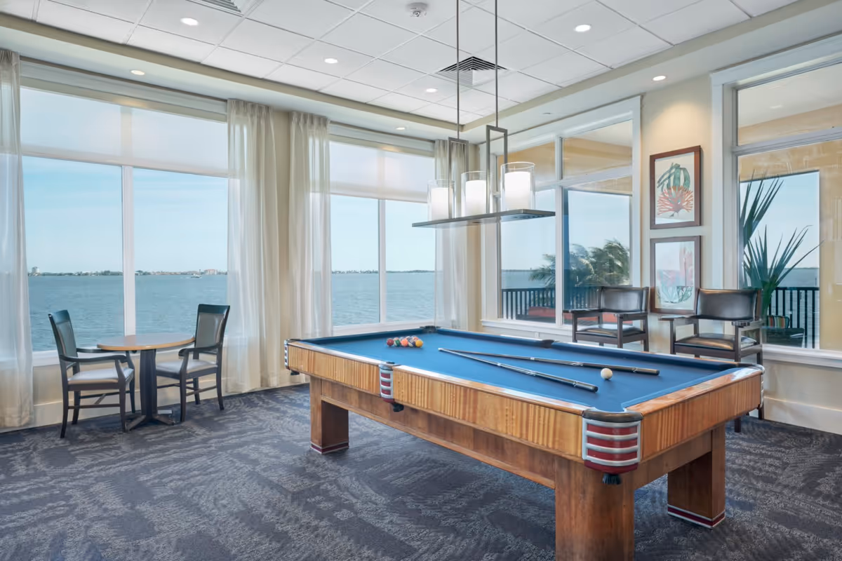 A bright room with large windows overlooking a body of water. The room features a pool table with cues and balls arranged on it, two chairs and a small round table near the windows, and two additional chairs against the wall with framed artwork above them. Light curtains hang beside the windows, and a modern light fixture is suspended above the pool table.