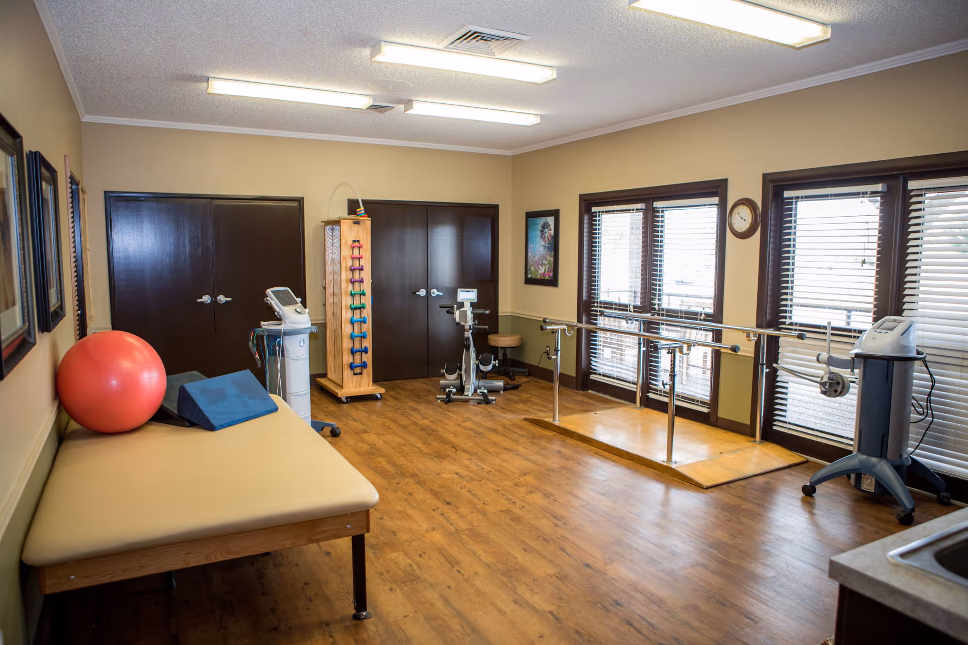 Physical therapy room with wooden flooring, a beige therapy table with a red exercise ball and blue wedge cushion, exercise equipment including a stationary bike, parallel bars for walking exercises, a rack of colorful dumbbells, and large windows with blinds letting in natural light.